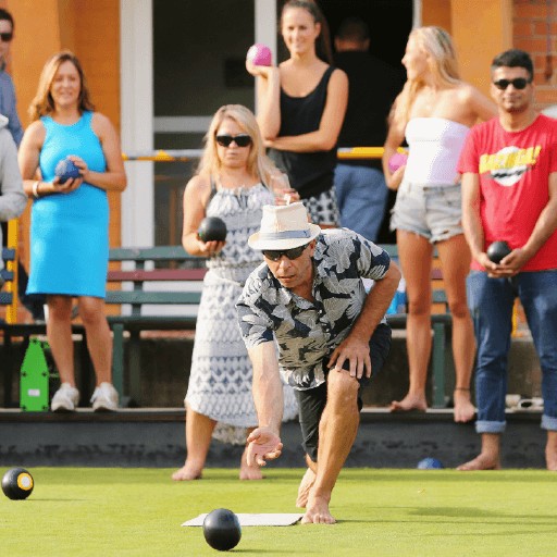 A middle aged man is doing barefoot lawn bowls in front of a crowd of onlookers